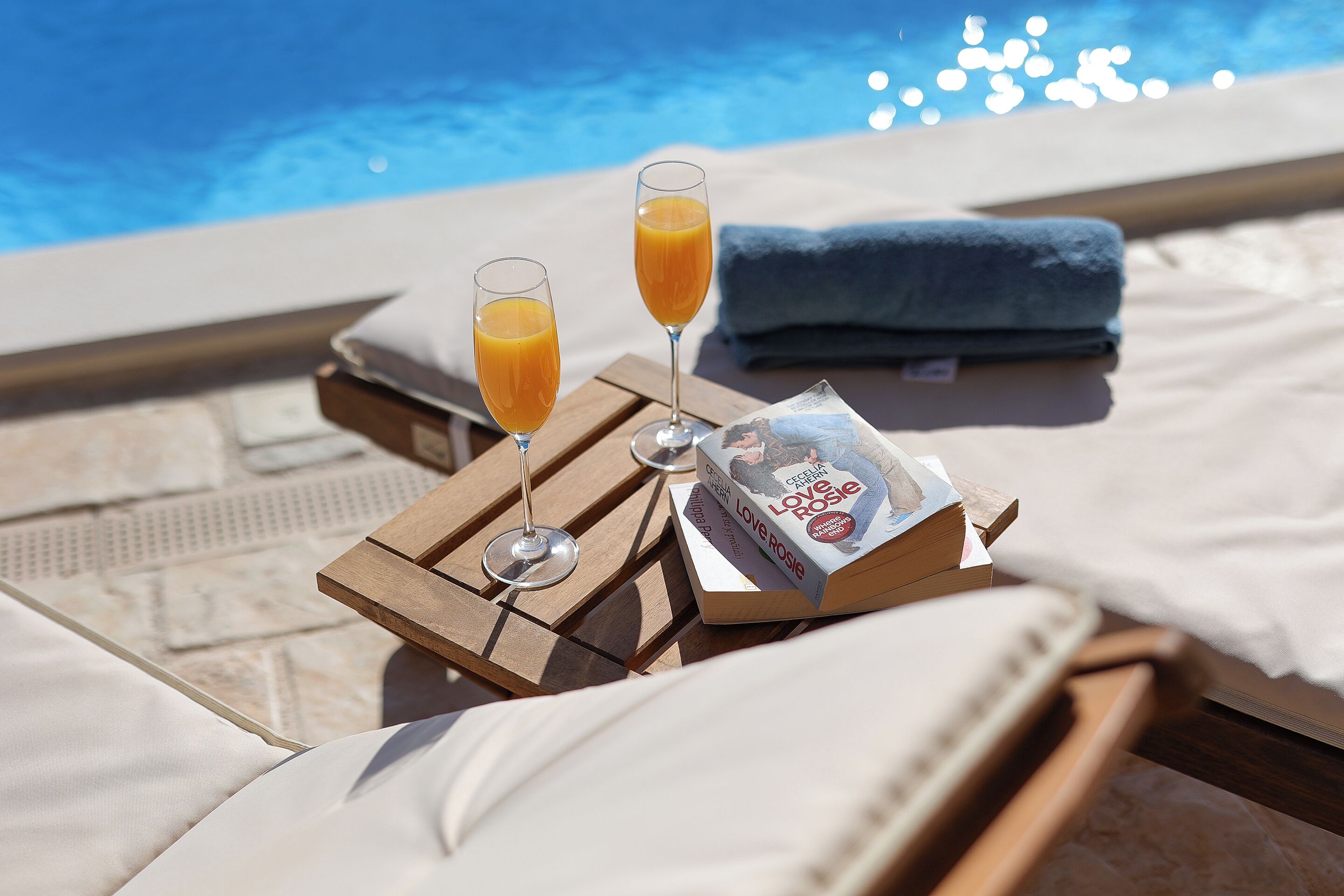 A poolside setting with lounge chairs, a tray with two glasses of orange juice, a book titled 'Love Rosie', and a towel.
