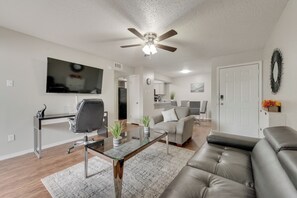 Een moderne woonkamer met een plafondventilator, een flatscreen-tv, een glazen salontafel, een leren bank en een eethoek met stoelen.