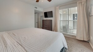 A bedroom with a bed, a ceiling fan, a TV, a window with blinds, and a door.