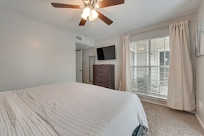 Un dormitorio con cama, ventilador de techo, televisión, cómoda y ventana con cortinas.