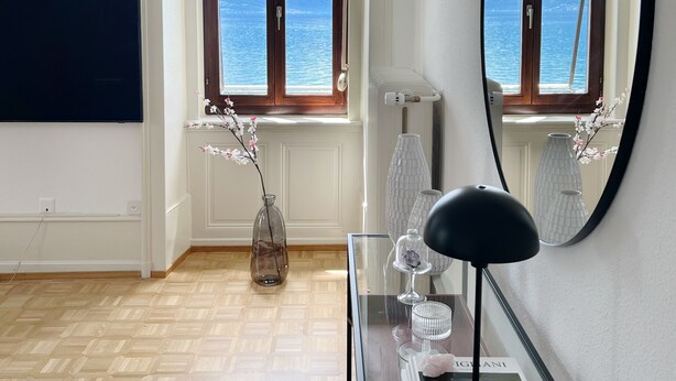 Una habitación con un ventanal grande, un espejo, una mesa de vidrio con libros y una lámpara, y un florero con flores.