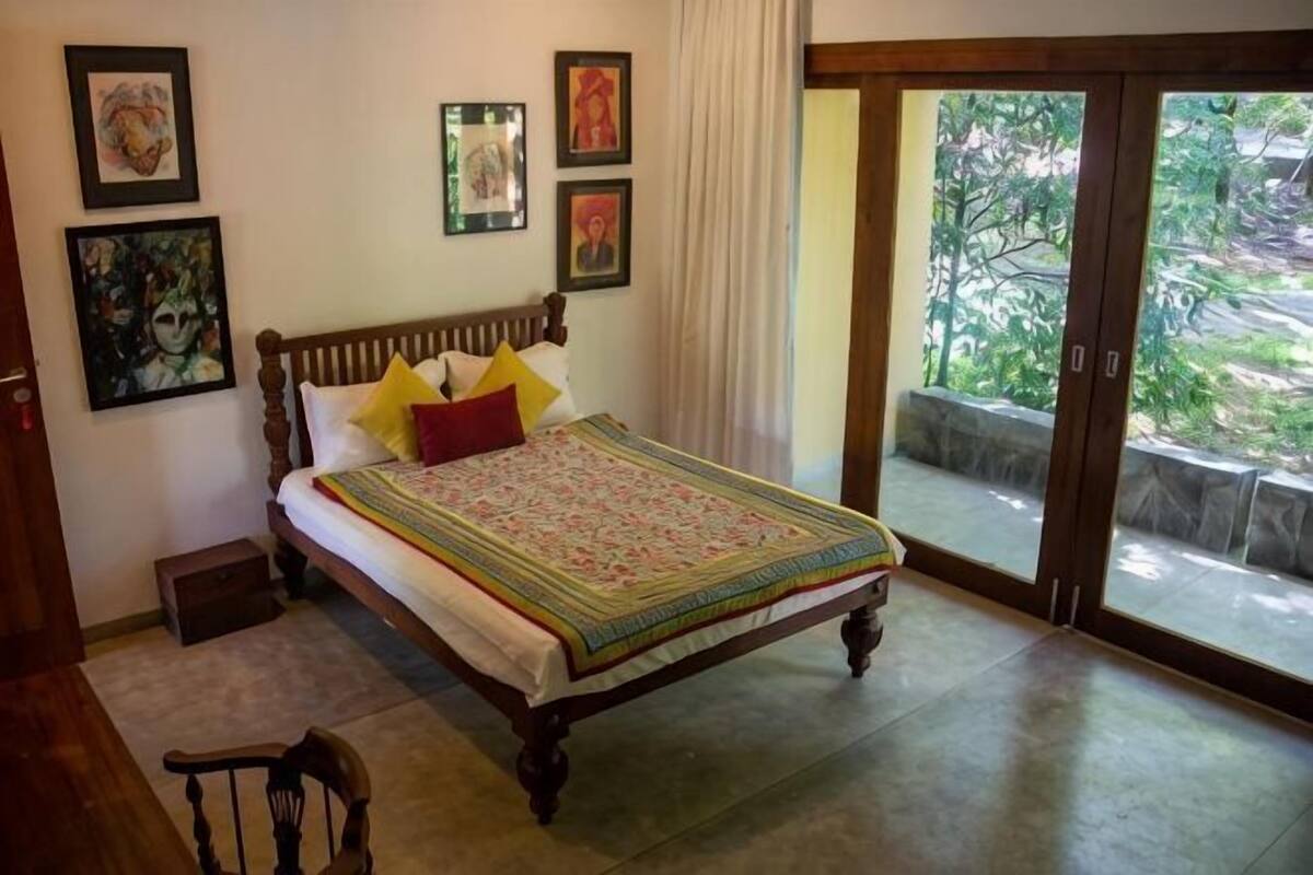 Ein Schlafzimmer mit einem hölzernen Bett, einem kleinen Nachttisch, einem Sessel und Blick ins Freie durch eine Schiebetür.