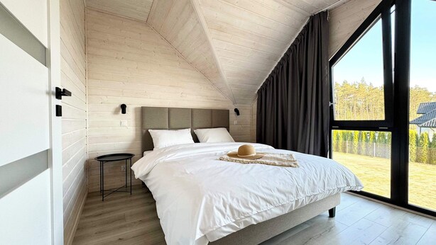 Une chambre moderne avec un plafond en pente, un grand lit avec une literie blanche, une petite table de chevet et une vue sur l’extérieur grâce à une grande fenêtre.