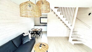 Un salon moderne avec un canapé gris foncé, deux tables basses en bois, des suspensions en osier, un coin repas avec une table ronde et des chaises, et un escalier blanc.