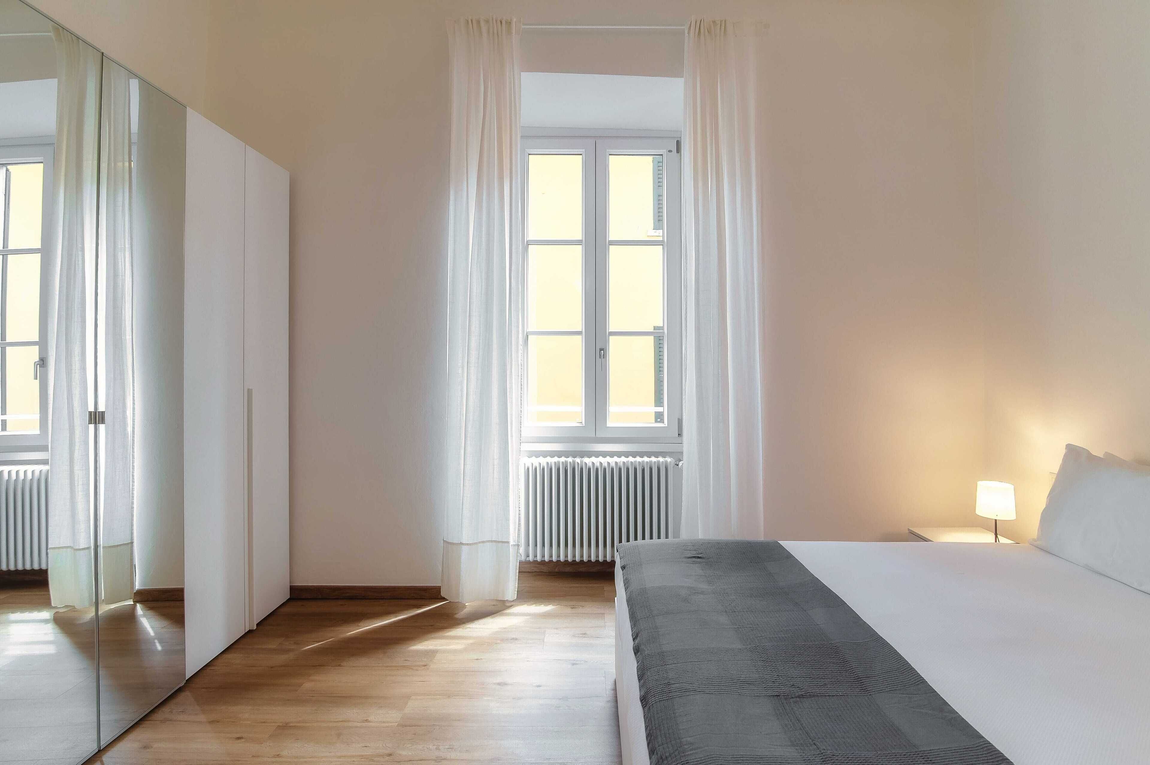 Una camera d'albergo con un letto, un comodino con una lampada, una finestra con tende, un termosifone e uno specchio a parete.