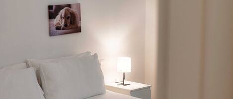 A neatly made bed with white linens, a bedside lamp, and two framed pictures of dogs on the wall.