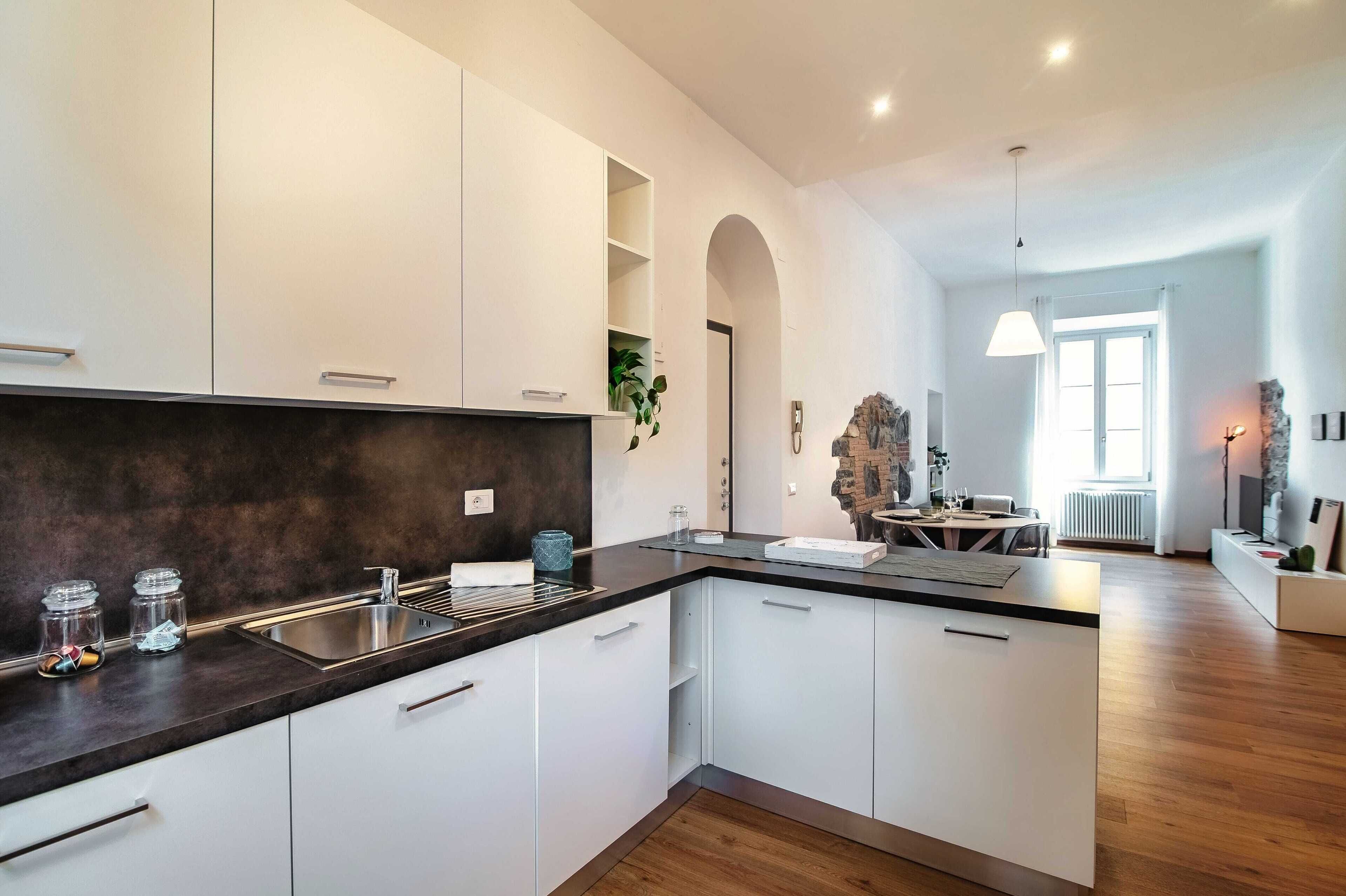 A modern kitchen with white cabinets, a dark countertop, and a built-in oven. There is a dining area with a table and chairs visible through an archway.