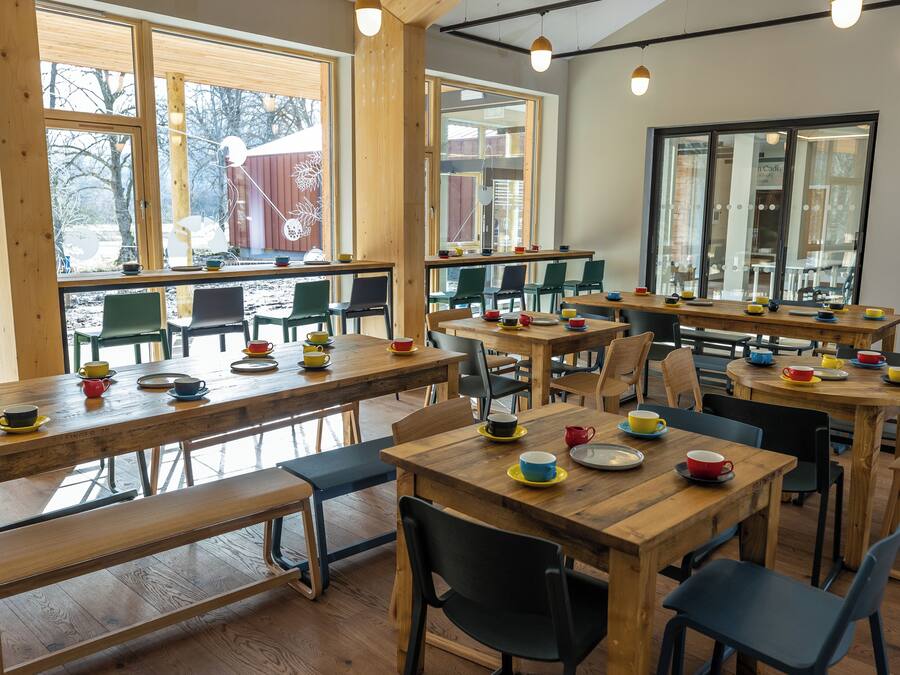 A spacious café with wooden tables and chairs, large windows, and a view of the outdoors.