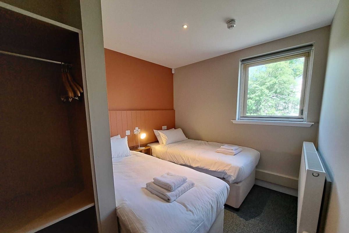 A hotel room with two beds, a wardrobe, a bedside lamp, and a window with a view of greenery.