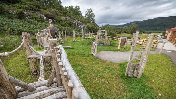 Un pont de bois rustique mène à une structure en bois surmontée d’un toit circulaire, le tout étant entouré d’une végétation luxuriante et avec des collines en arrière-plan.