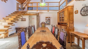 Un espace repas comprenant une longue table en bois, des chaises décorées, un escalier et un buffet en bois doté de portes en verre.