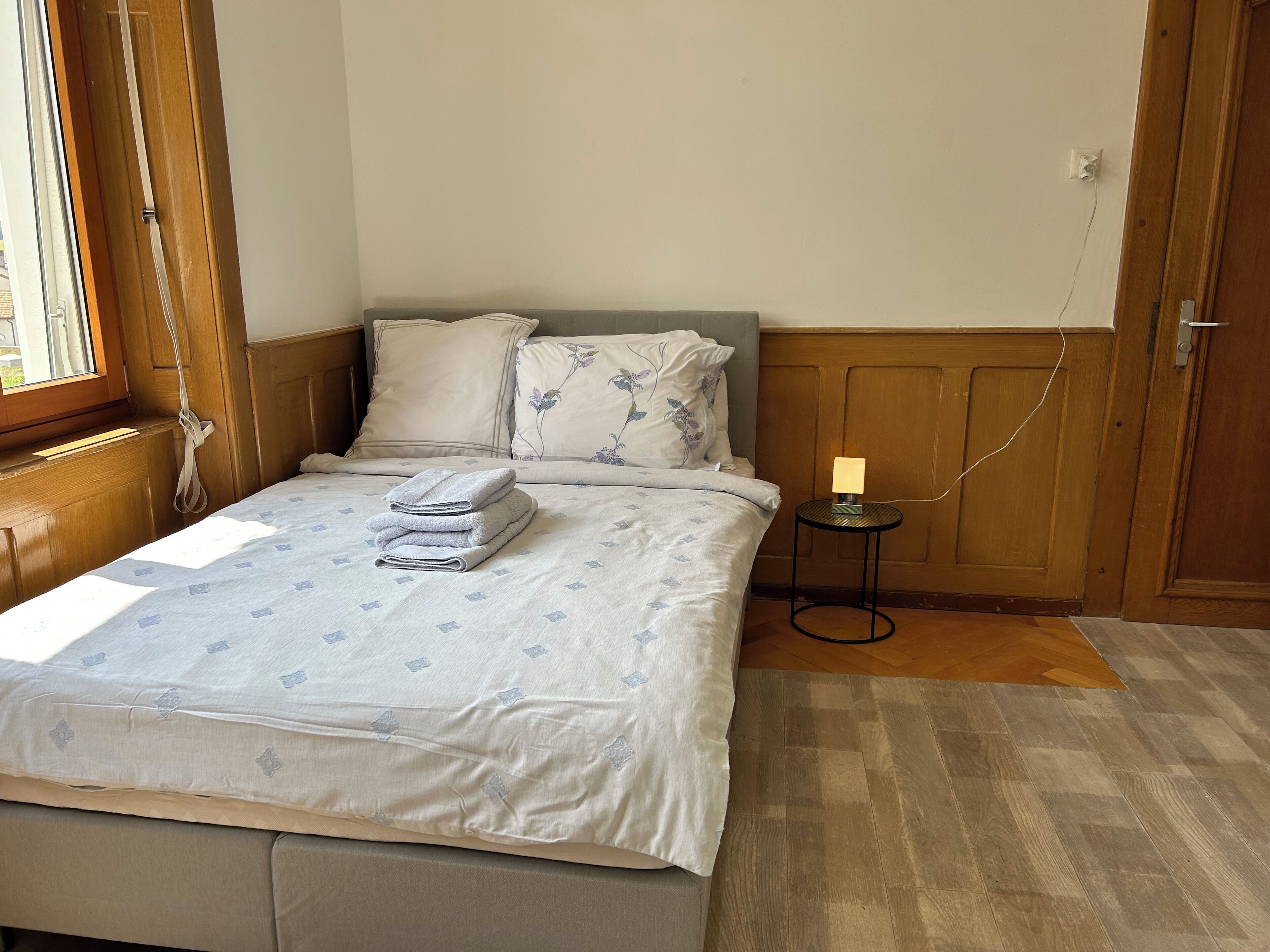 Ein ordentlich bezogenes Bett mit einem gemusterten Überwurf, ein Stapel gefalteter Handtücher und eine Nachttischlampe auf einem kleinen Tisch.