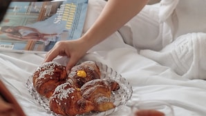 Una persona asseguda a un llit, amb un got de beguda d'aranja, aguantant una revista i un plat de pastissos.
