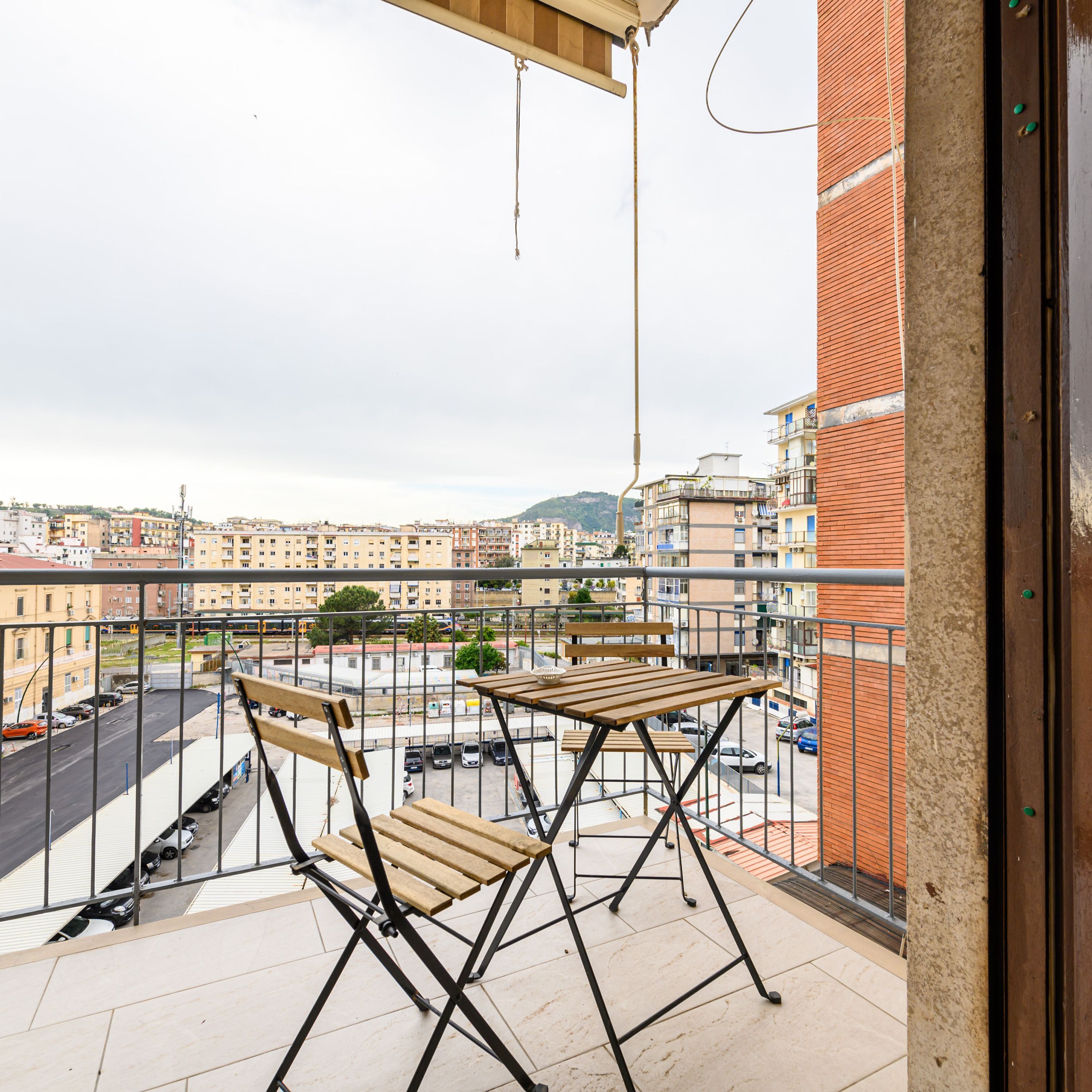 A balcony with a wooden table and chairs, overlooking a cityscape with buildings and a road.