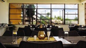 Un restaurante con varias mesas, sillas negras y un amplio ventanal con vistas a una escalera y follaje.