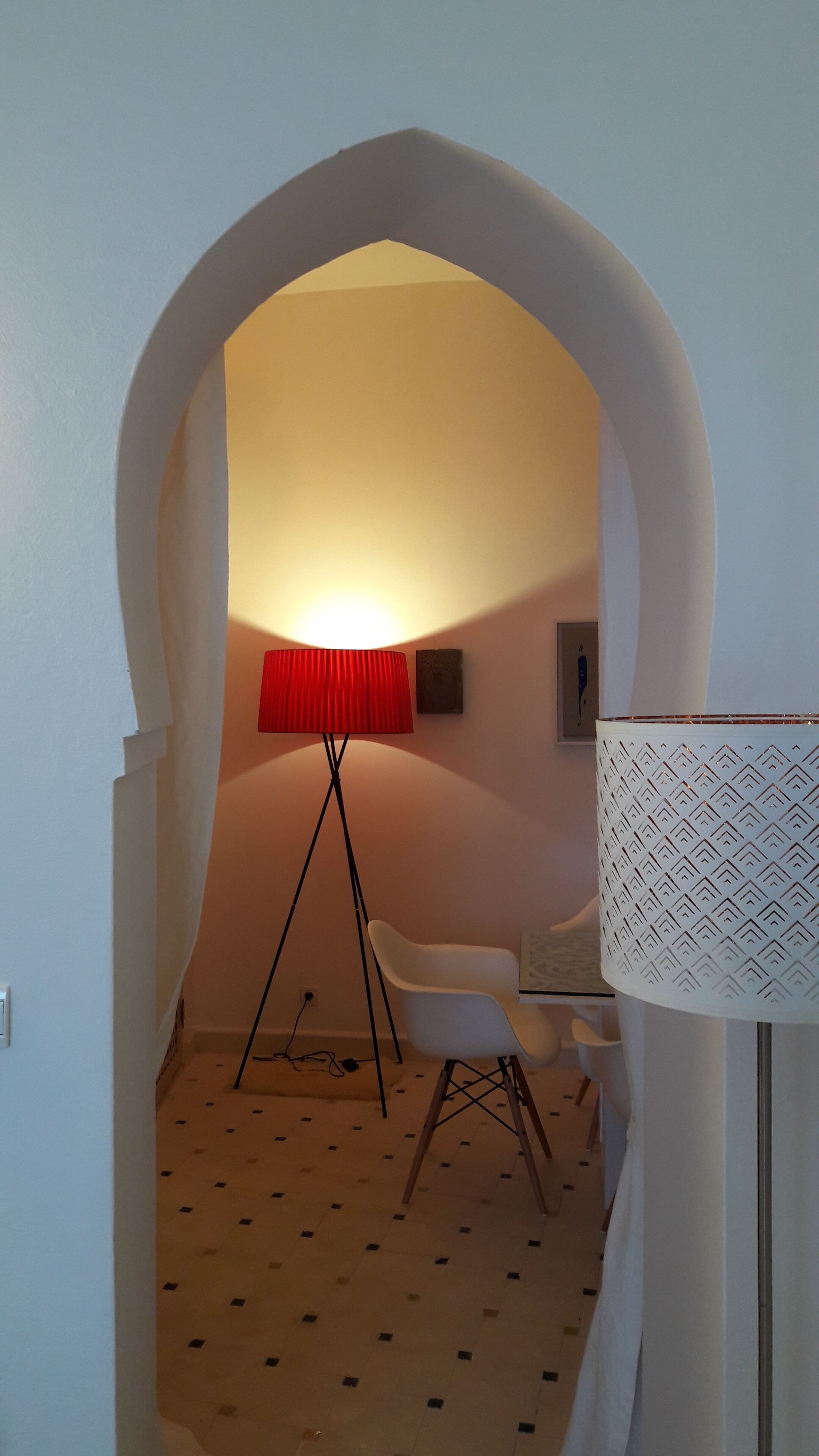 Um corredor estreito com uma porta em arco que dá para um quarto. Uma luminária vermelha no chão e uma luminária branca com cúpula estampada são visíveis. Uma cadeira branca e uma pequena mesa estão no quarto.