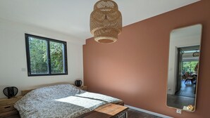 Une chambre à coucher avec un grand lit, une table de chevet en bois, une commode, un grand miroir et une fenêtre donnant sur un paysage verdoyant.