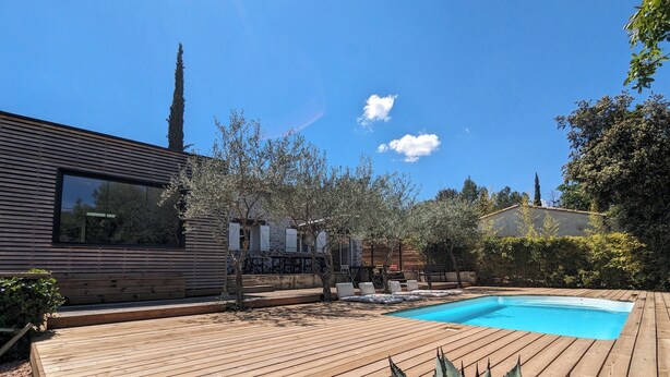 Une terrasse en bois avec une piscine, entourée d’arbres et bordée d’un bâtiment moderne.
