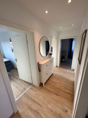 A hallway with wooden flooring, a white door, a white dresser with drawers, a round mirror, and recessed ceiling lighting.