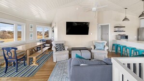 A spacious living area with a wooden dining table, blue chairs, a grey sofa, and a kitchen area with a microwave and stools. - Block Island - 4 Bedroom Home on West Beach Road w/ Panoramic Ocean Views + AC! (New Shoreham)