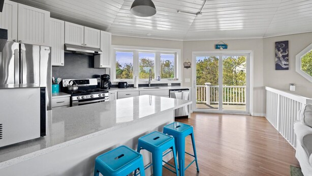 A modern kitchen with white cabinetry, stainless steel appliances, and a large island with blue stools. - Block Island - 4 Bedroom Home on West Beach Road w/ Panoramic Ocean Views + AC! (New Shoreham)
