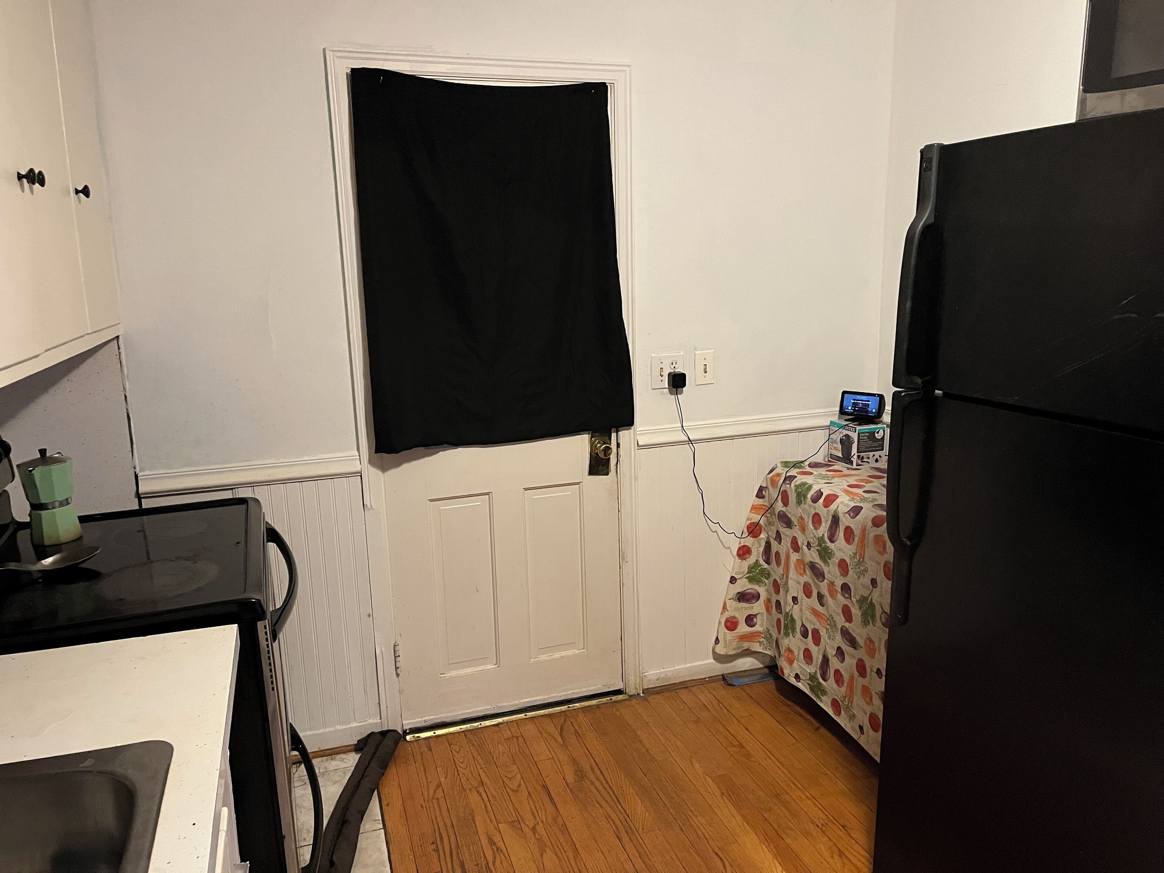 Une cuisine compacte équipée d’un réfrigérateur, d’un four à micro-ondes et d’une petite table recouverte d’une nappe à motifs de fruits.