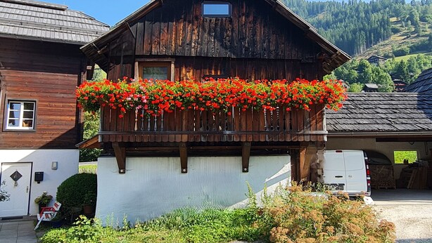 A wooden chalet with a balcony adorned with red flowers, a white door, and a small window. - Die Alte Scheune by Globalimmoservice (Bad Kleinkirchheim)