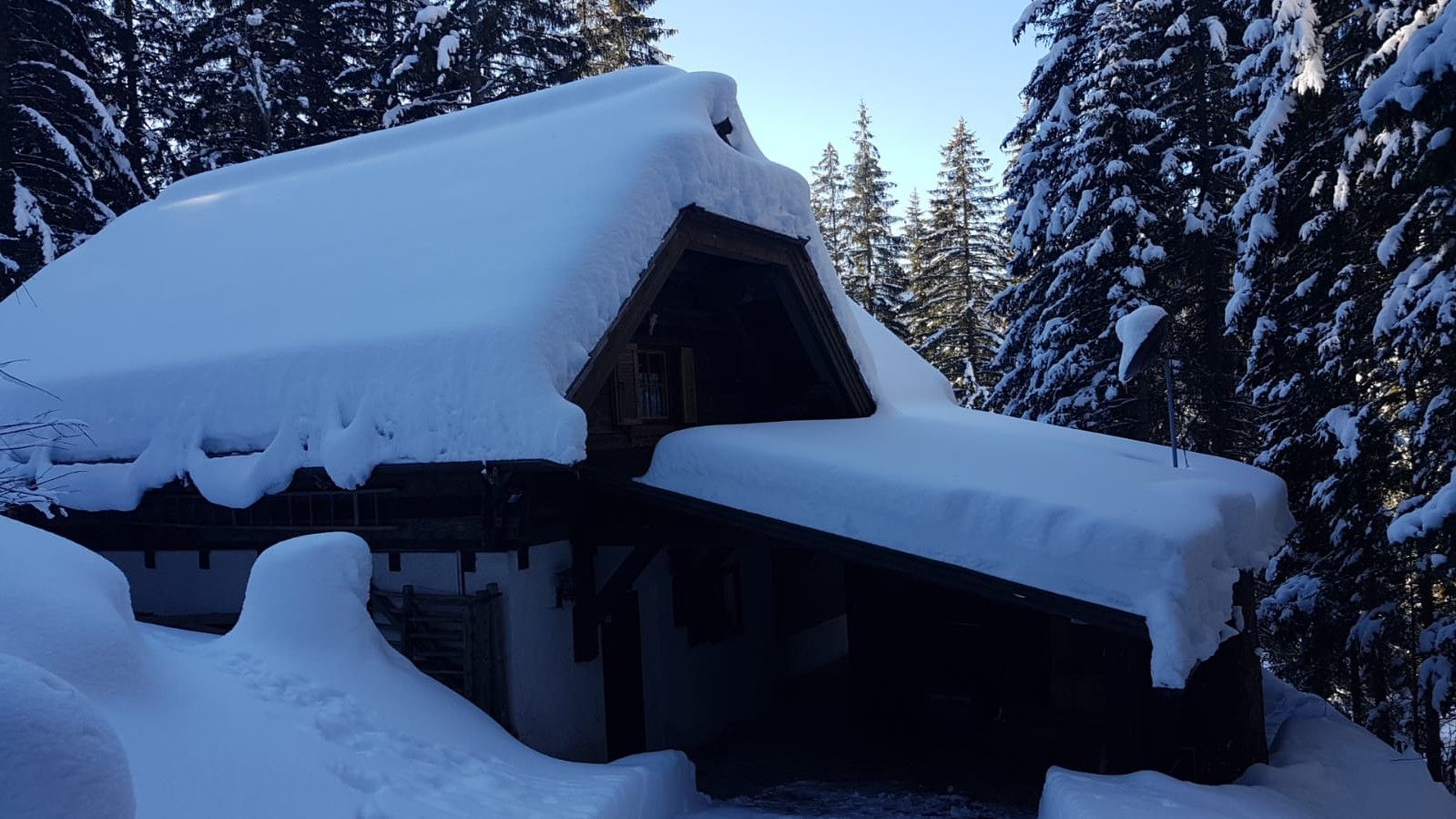 Une cabane en bois recouverte de neige, avec un toit à forte pente, entourée d’épais conifères.