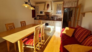 Ein gemütliches Wohnzimmer mit einem Holzesstisch, rot gestreiften Stühlen und einem roten Sofa mit gelben Kissen. Im Hintergrund ist eine Küche mit weißen Schränken und einer Mikrowelle zu sehen.