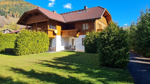 A two-story wooden chalet with a sloped roof, a balcony, and a well-maintained lawn.