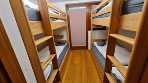 Ein schmales Zimmer mit in U-Form angeordneten Etagenbetten, Holzboden und einem Fenster mit Jalousien.