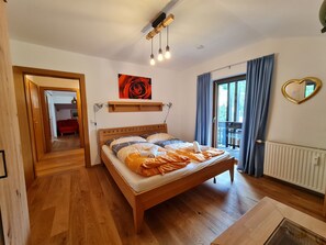 A bedroom with a wooden bed, a red sofa in the adjacent room, a wall-mounted shelf, a heart-shaped mirror, and a window with blue curtains.