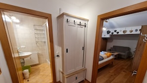 Un corridoio con pavimento in legno, un armadio bianco con ganci, un bagno con lavandino e doccia, e una zona living con divano e tavolo.