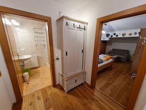 Un pasillo con piso de madera, un armario blanco con ganchos, un baño con lavamanos y ducha, y una sala de estar con sofá y mesa.