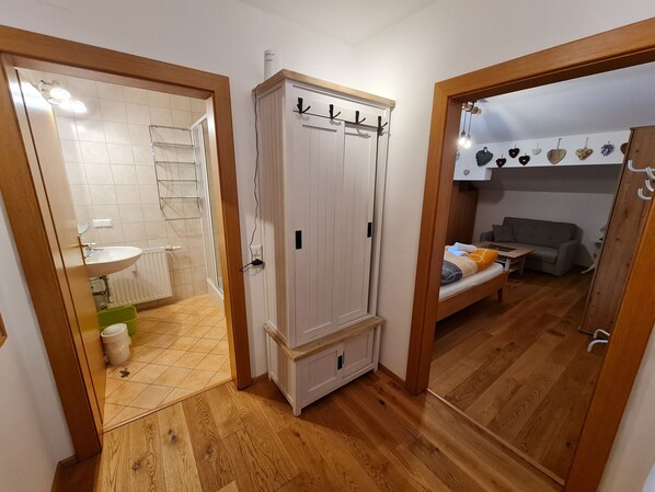 A hallway with a wooden floor, a white wardrobe with hooks, a bathroom with a sink and a shower, and a living area with a sofa and a table.