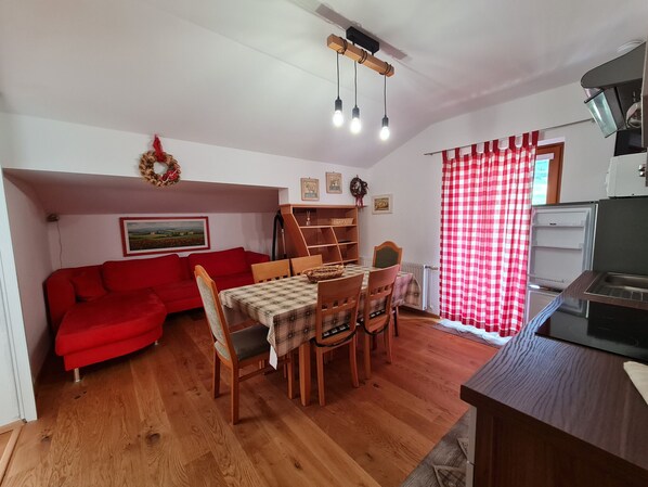A compact living space with a red sofa, wooden dining table, and chairs. A kitchen area with a refrigerator and a microwave is visible.
