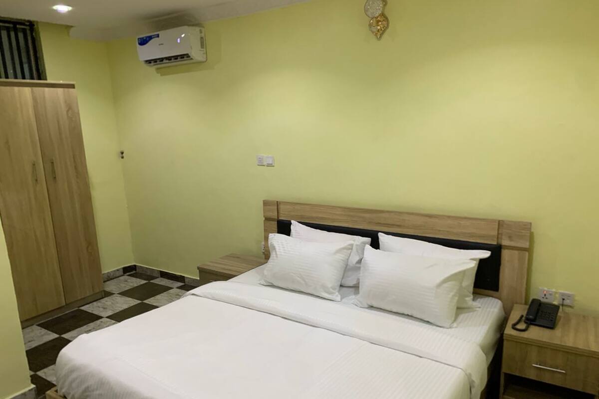 A hotel room with a bed, a nightstand, a wardrobe, and an air conditioning unit on the wall.
