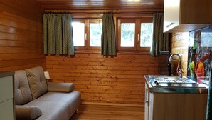 A cozy cabin interior with a wooden floor, a sofa, a kitchen area with a sink and stove, and windows with patterned curtains. - Der Kristall Top 3 by Globalimmoservice (Bad Kleinkirchheim)