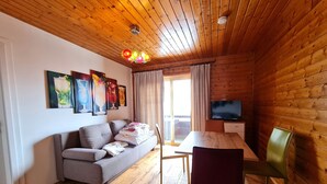 A cozy cabin-style room with a wooden ceiling, a sofa, a dining table with chairs, and a TV mounted on the wall.