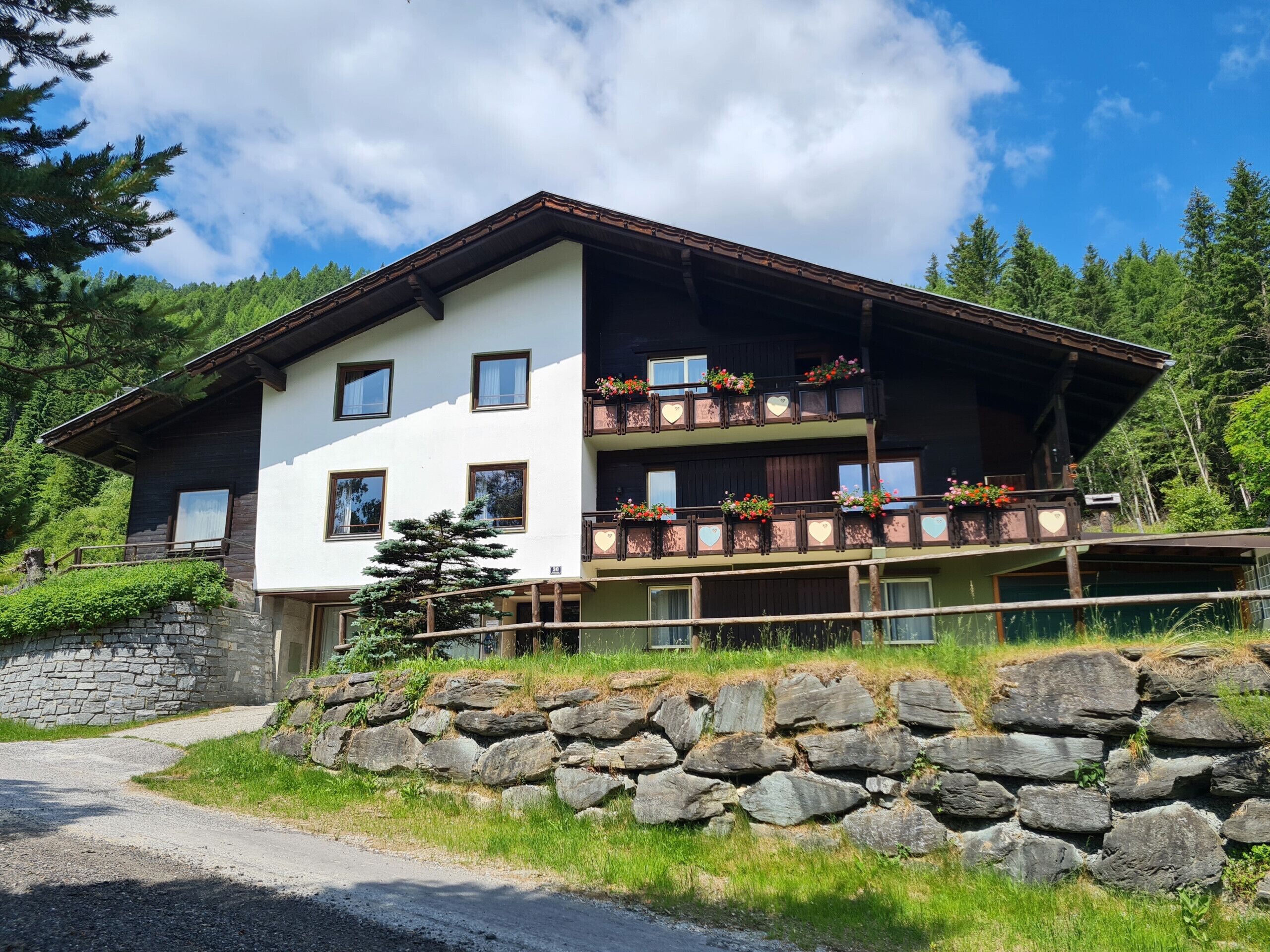 A two-story chalet with a stone foundation, wooden balconies with flower boxes, and a clear view of the surrounding forest.
