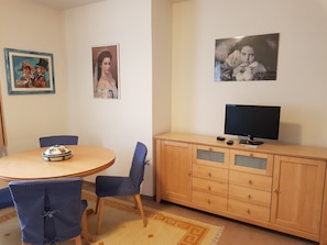 Una habitación con una mesa de comedor redonda, sillas azules, un mueble de madera con un televisor y cuadros en la pared.