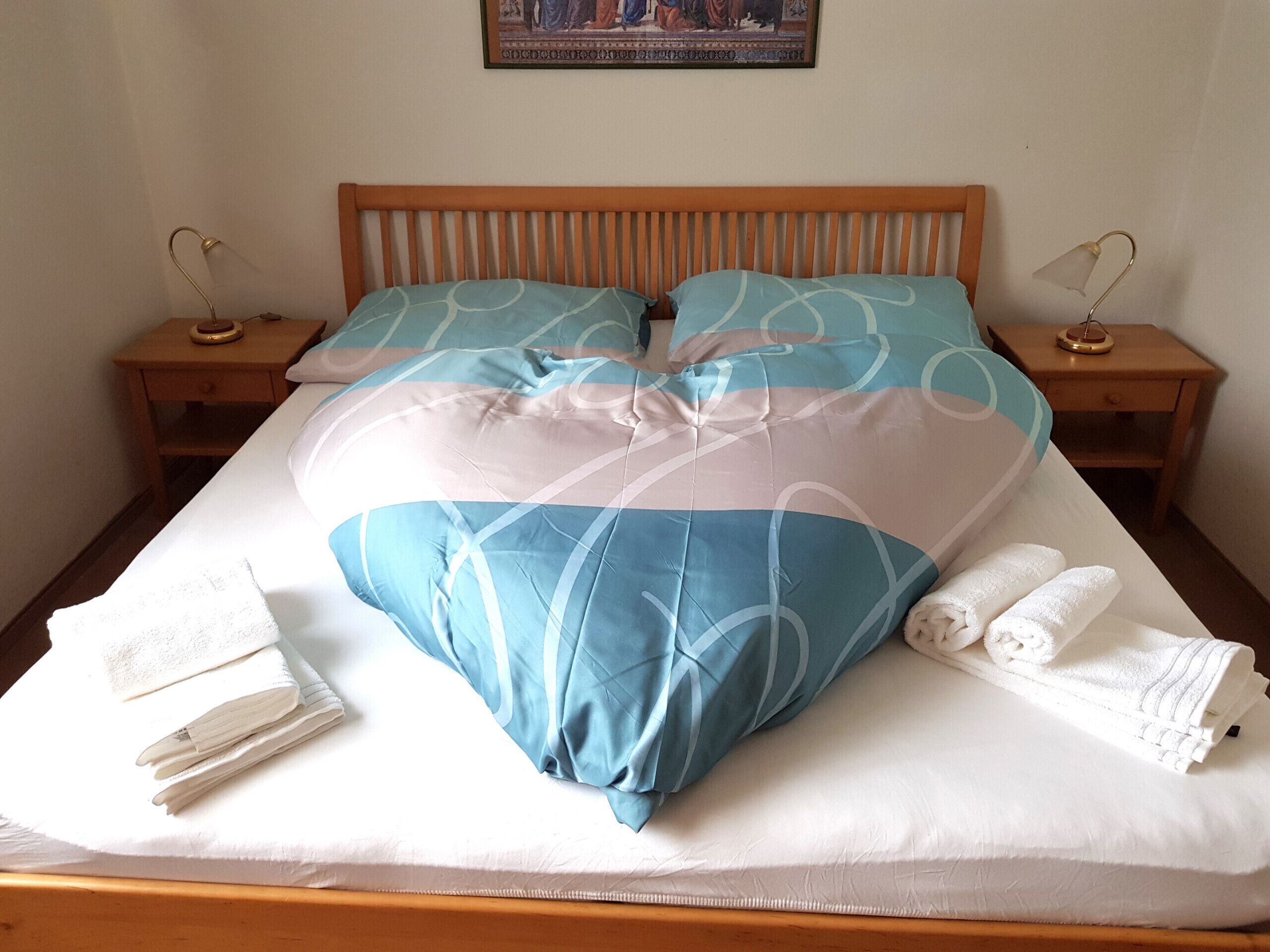 Una cama bien tendida con edredón azul y blanco a cuadros, dos mesitas de noche de madera con lámparas, y un cuadro en la pared.