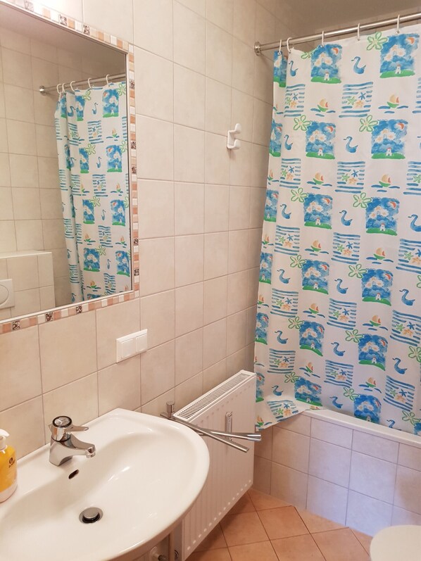 Un baño con lavamanos blanco, espejo, cortina de ducha con motivos náuticos y suelo de baldosas.