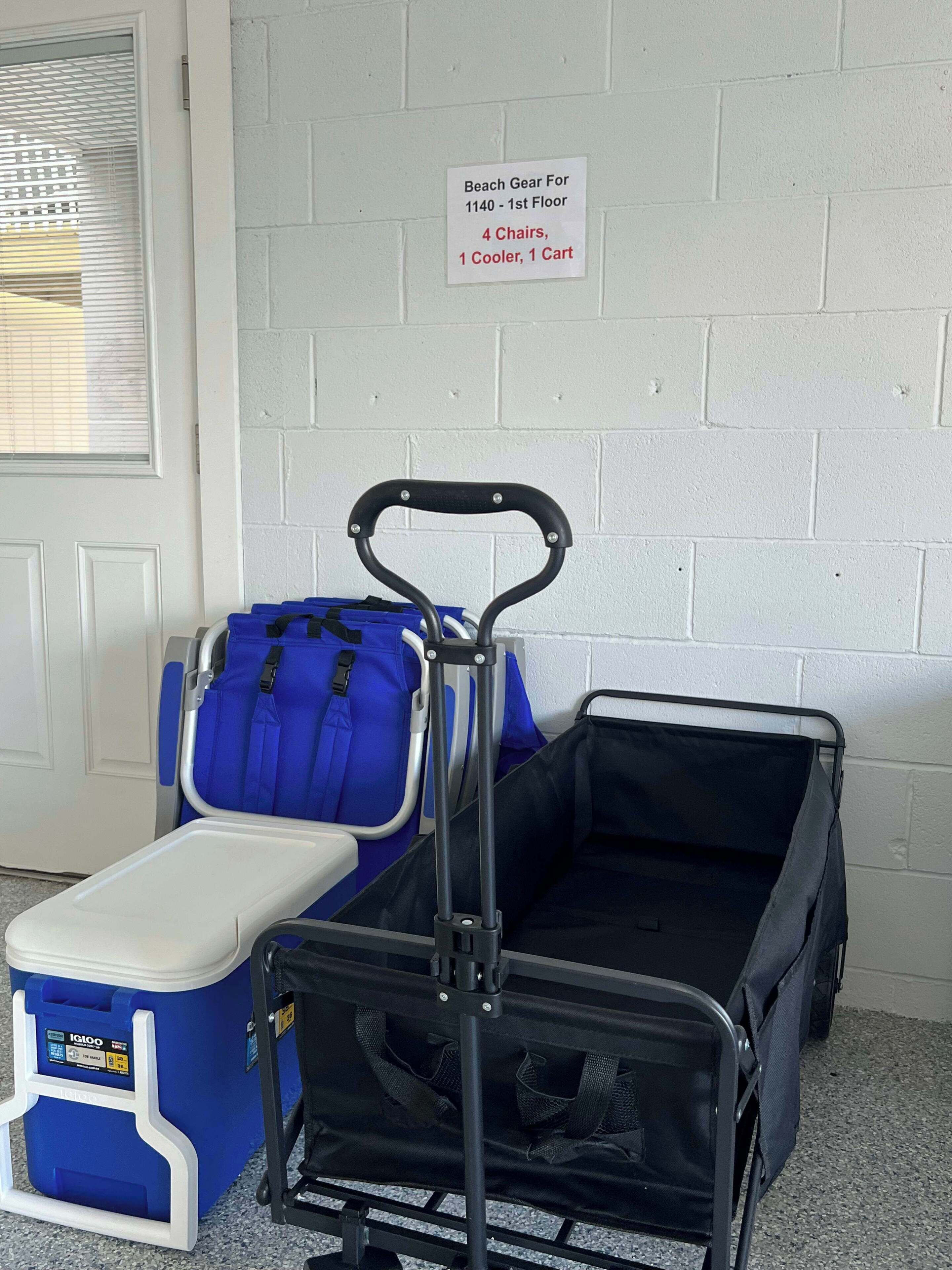 Carrinho com um cooler, uma bolsa azul e uma bolsa preta, com placa indicando equipamentos de praia para o 1º andar.