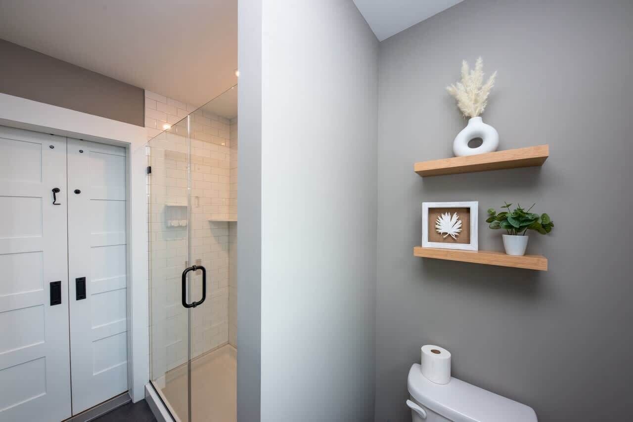 A modern bathroom with a glass shower enclosure, white toilet, and decorative shelves with plants and framed items.