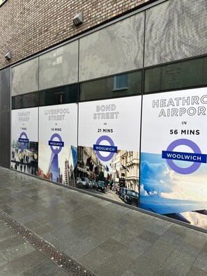 A building facade with advertisements for Canary Wharf, Liverpool Street, Bond Street, and Heathrow Airport, indicating travel times to Woolwich. - Stylish Elegant New Flat 5 Min from Crossrail (London)
