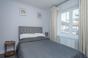 A bedroom with a grey upholstered bed, a round wooden side table, two framed abstract artworks on the wall, and a window with curtains. - Stylish Elegant New Flat 5 Min from Crossrail (London)