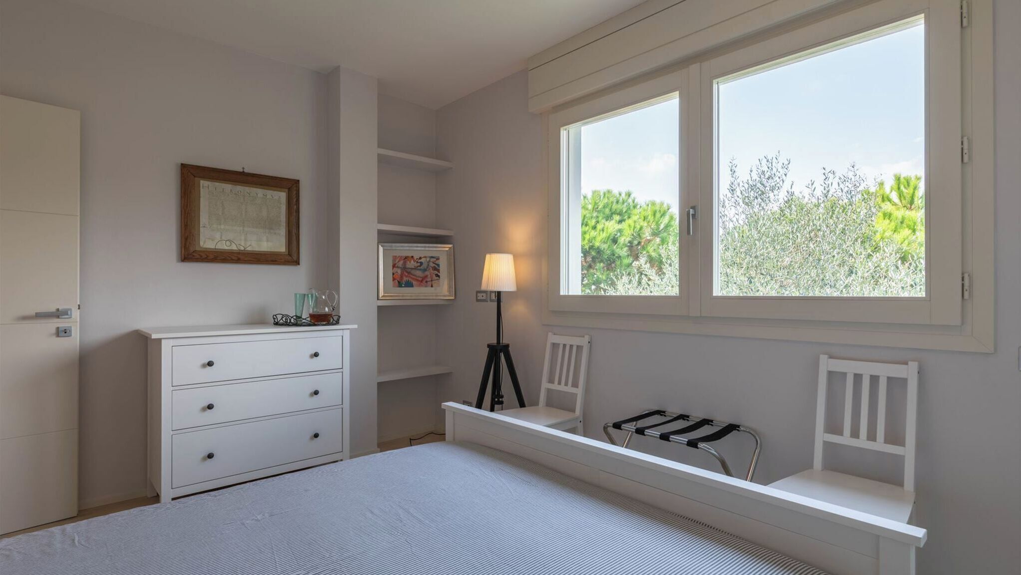 A bedroom with a bed, a dresser, a lamp, a window with a view of greenery, and a radiator.