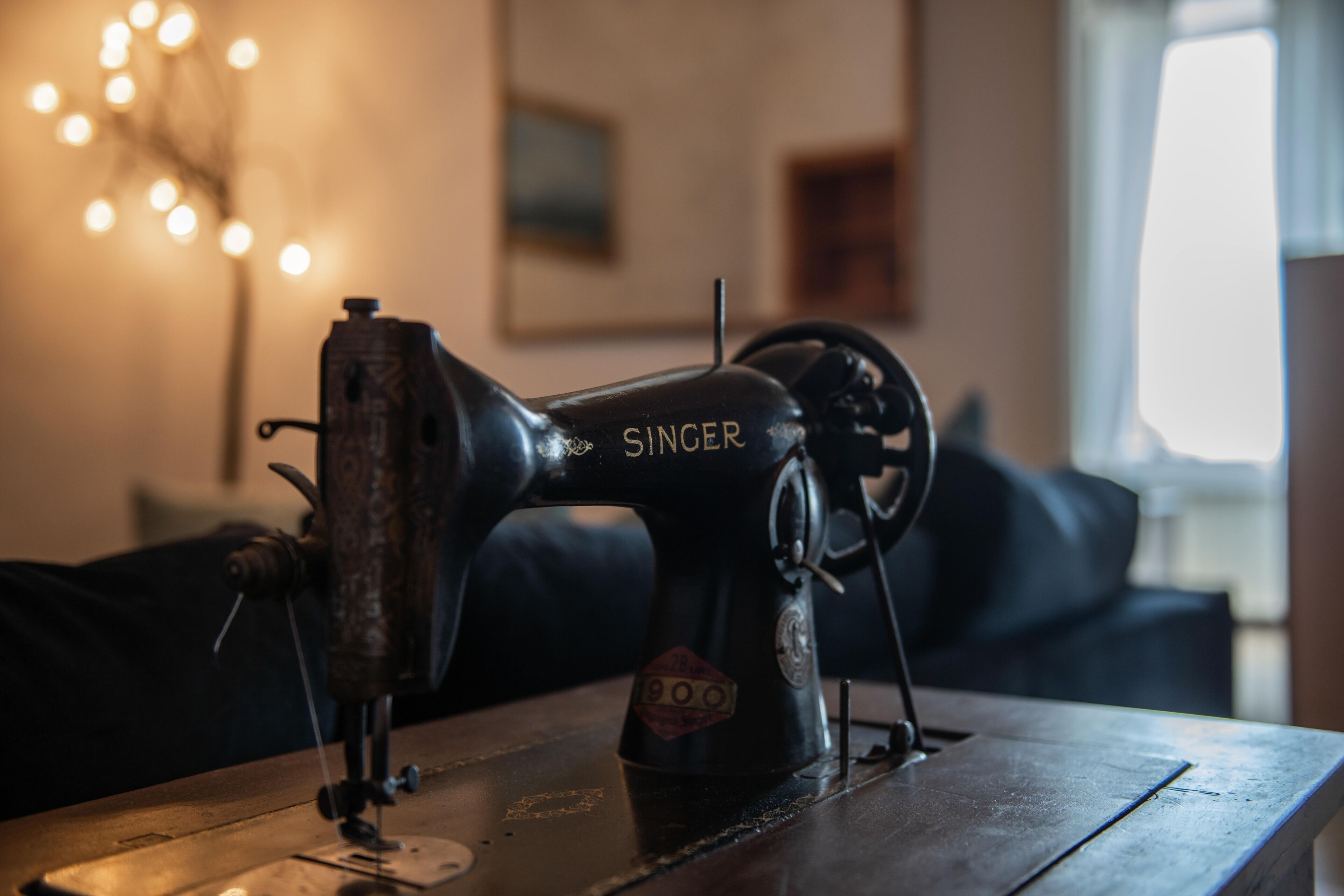 A Singer sewing machine on a wooden table in a room with a dark-colored sofa and a lamp with multiple bulbs in the background.