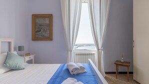 A bedroom with a bed, a nightstand, a lamp, a window with curtains, and a painting on the wall. - Italianway - Vele D'Amare (Imperia)